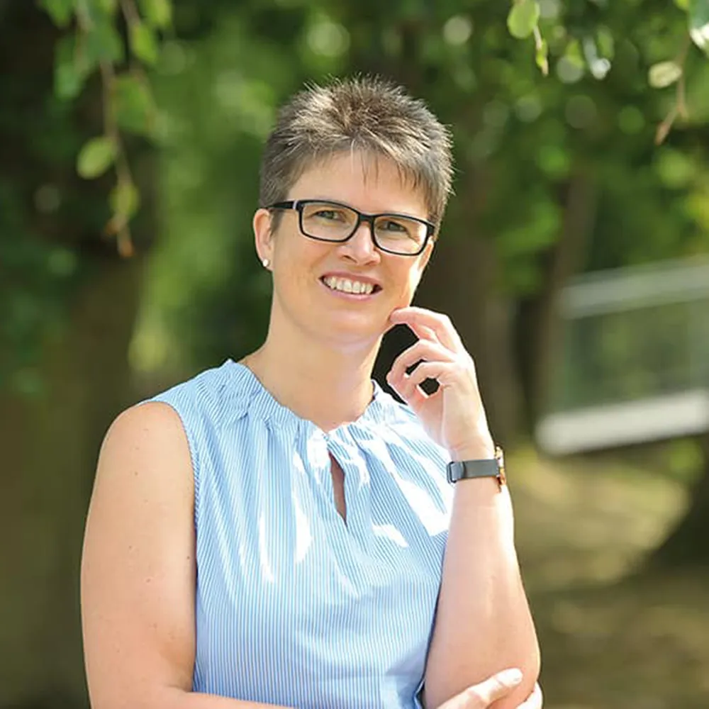 Eine Person mit kurzen Haaren und Brille lächelt im Freien, sie trägt ein ärmelloses blaues Oberteil und eine Uhr, im Hintergrund sind Bäume und Grünflächen zu sehen.