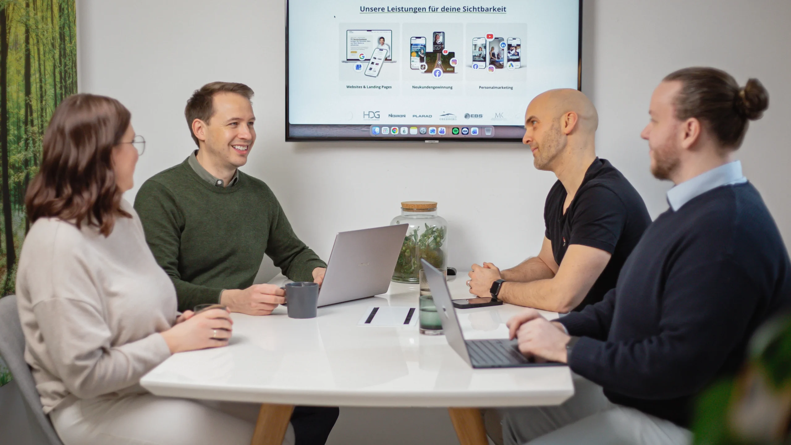 Vier Personen sitzen mit Laptops und Kaffeetassen um einen weißen Tisch und diskutieren. Auf einem Bildschirm im Hintergrund ist eine Präsentationsfolie zu sehen.