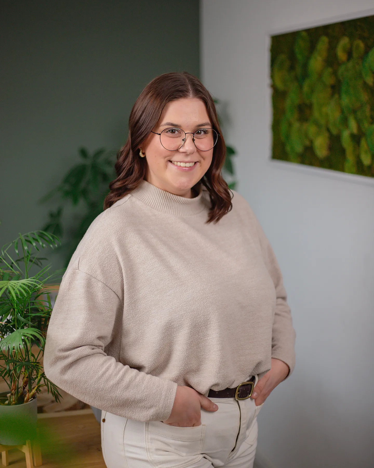 Eine Frau mit Brille, die einen beigen Pullover und eine weiße Hose trägt, steht in einem Innenraum mit Pflanzen und einer grünen Wandmalerei im Hintergrund und lächelt in die Kamera.