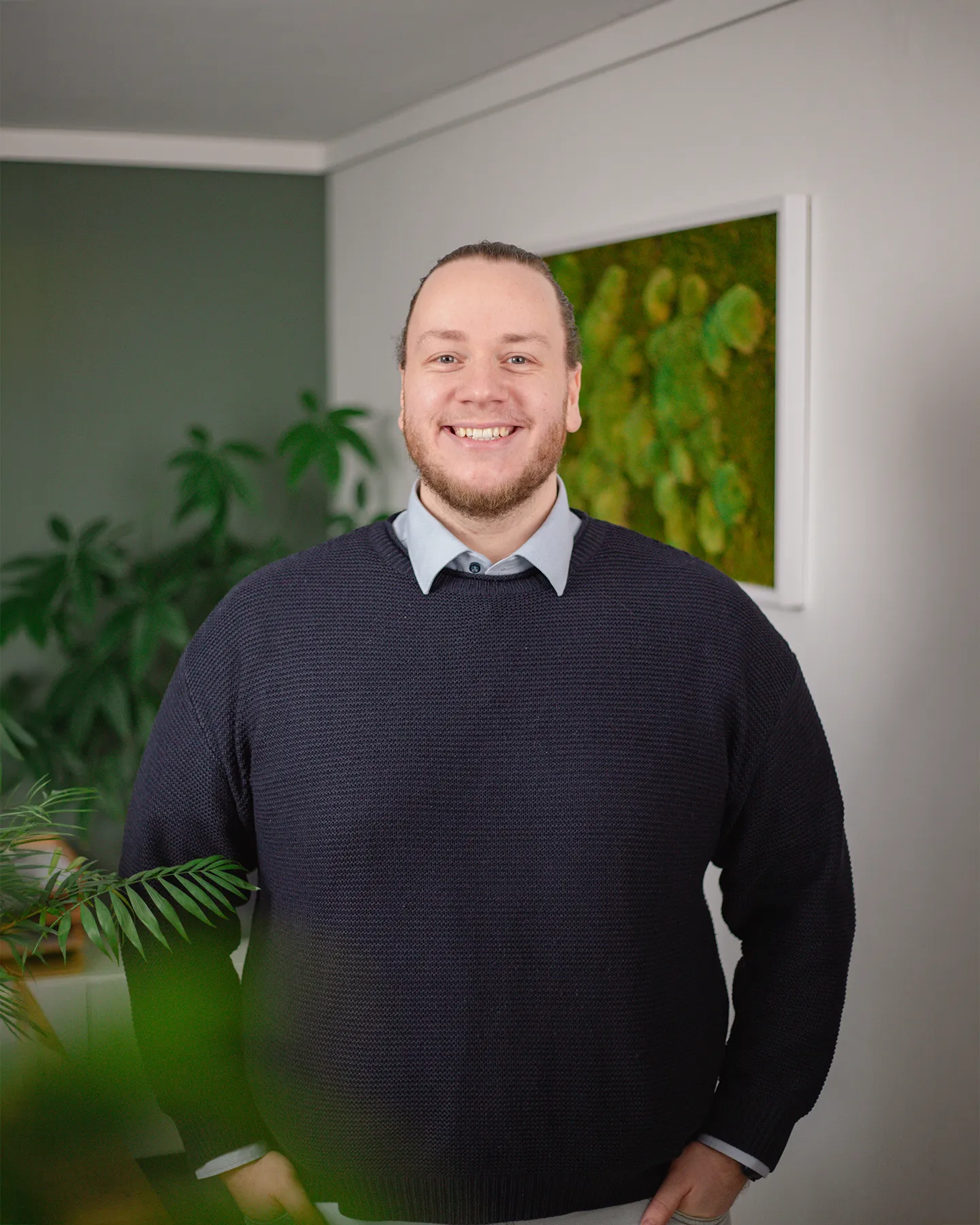 Ein Mann mit Bart, der einen marineblauen Pullover über einem Kragenhemd trägt, steht lächelnd in einem Raum mit Grünpflanzen und einem gerahmten Bild an der Wand.
