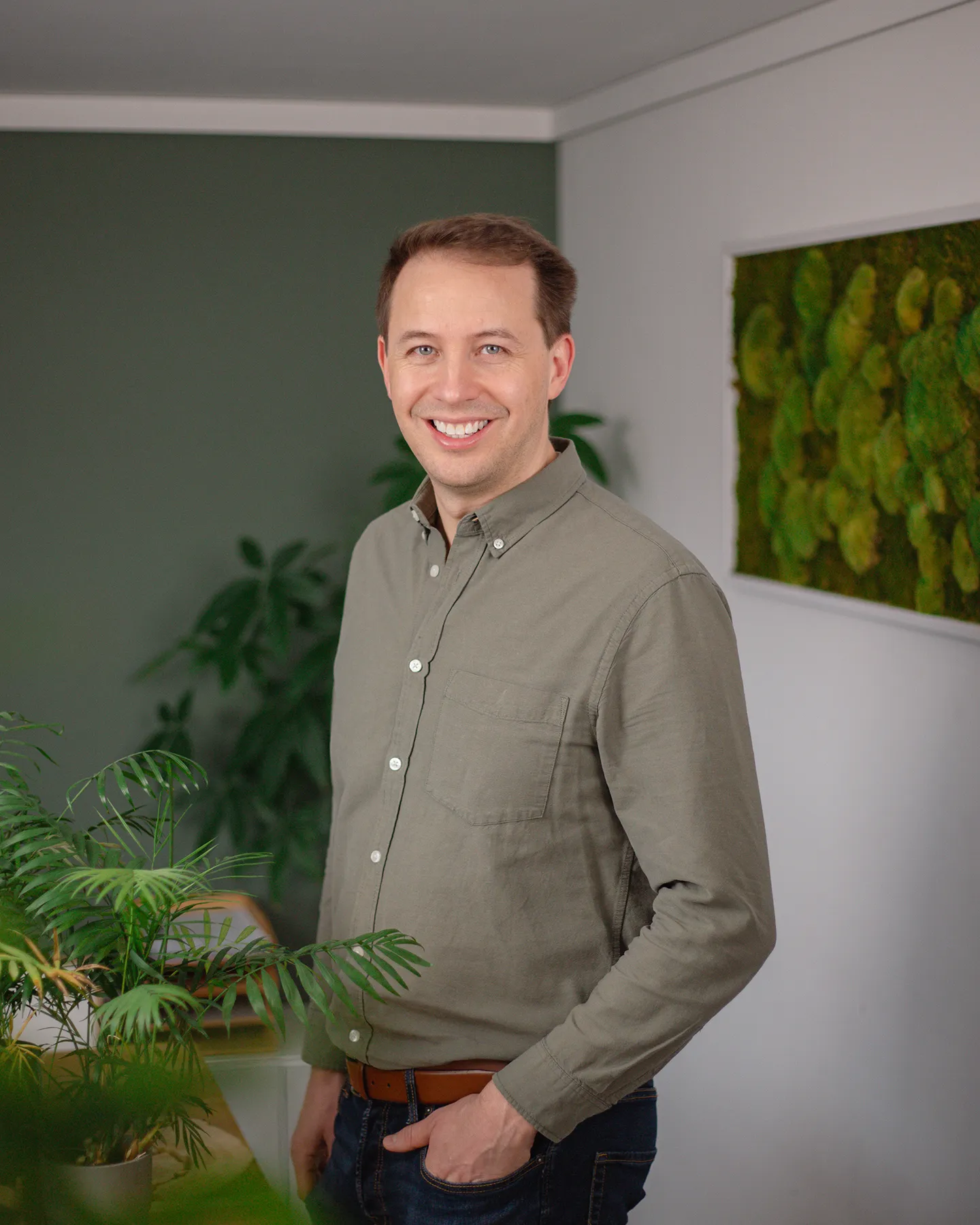 Ein Mann in einem grauen Button-up-Hemd steht in einem Haus und lächelt in die Kamera. Im Hintergrund sind Grünpflanzen und eine grüne Wanddekoration zu sehen.