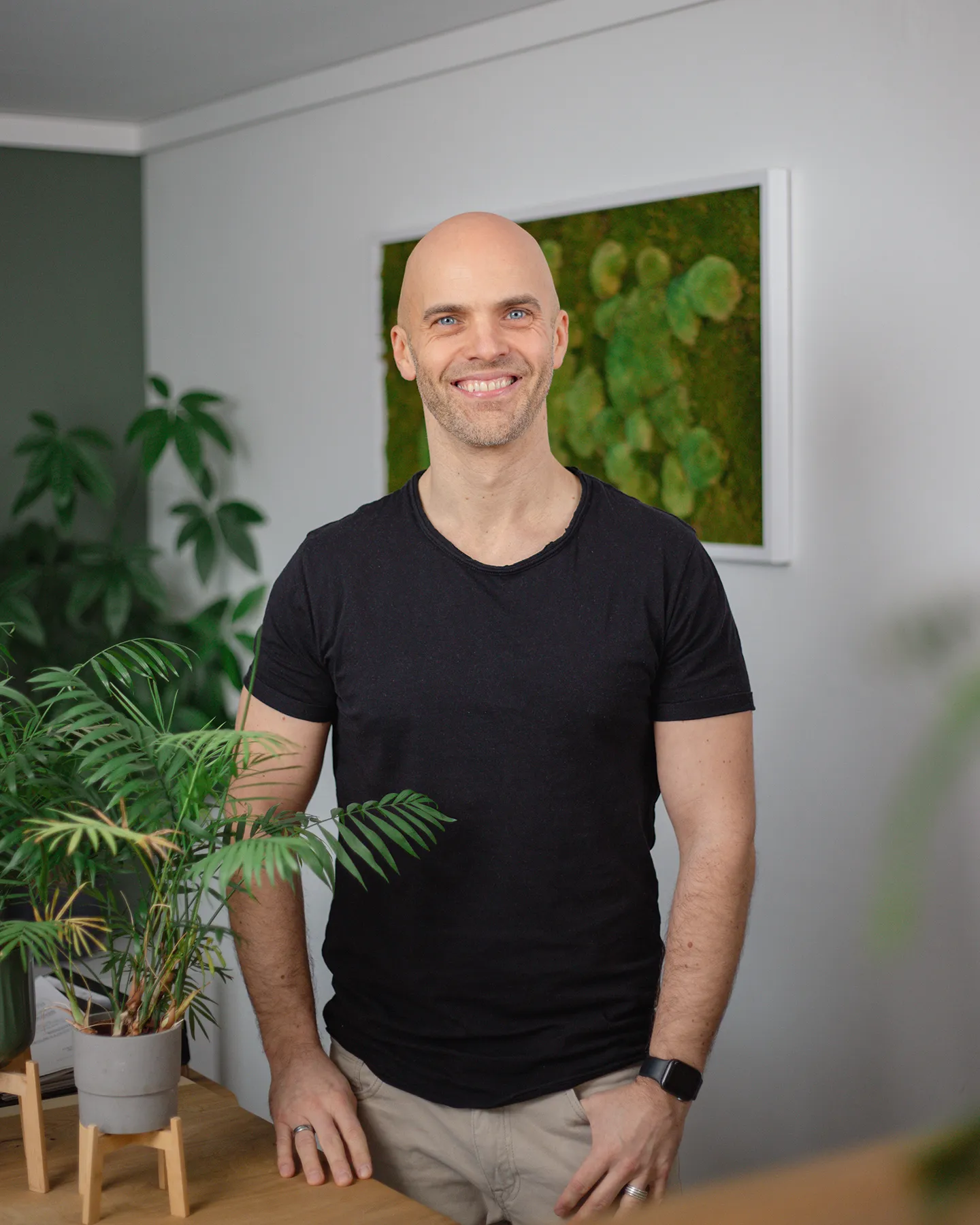 Ein Mann mit rasiertem Kopf und schwarzem T-Shirt steht in einem Haus neben grünen Topfpflanzen und lächelt in die Kamera.