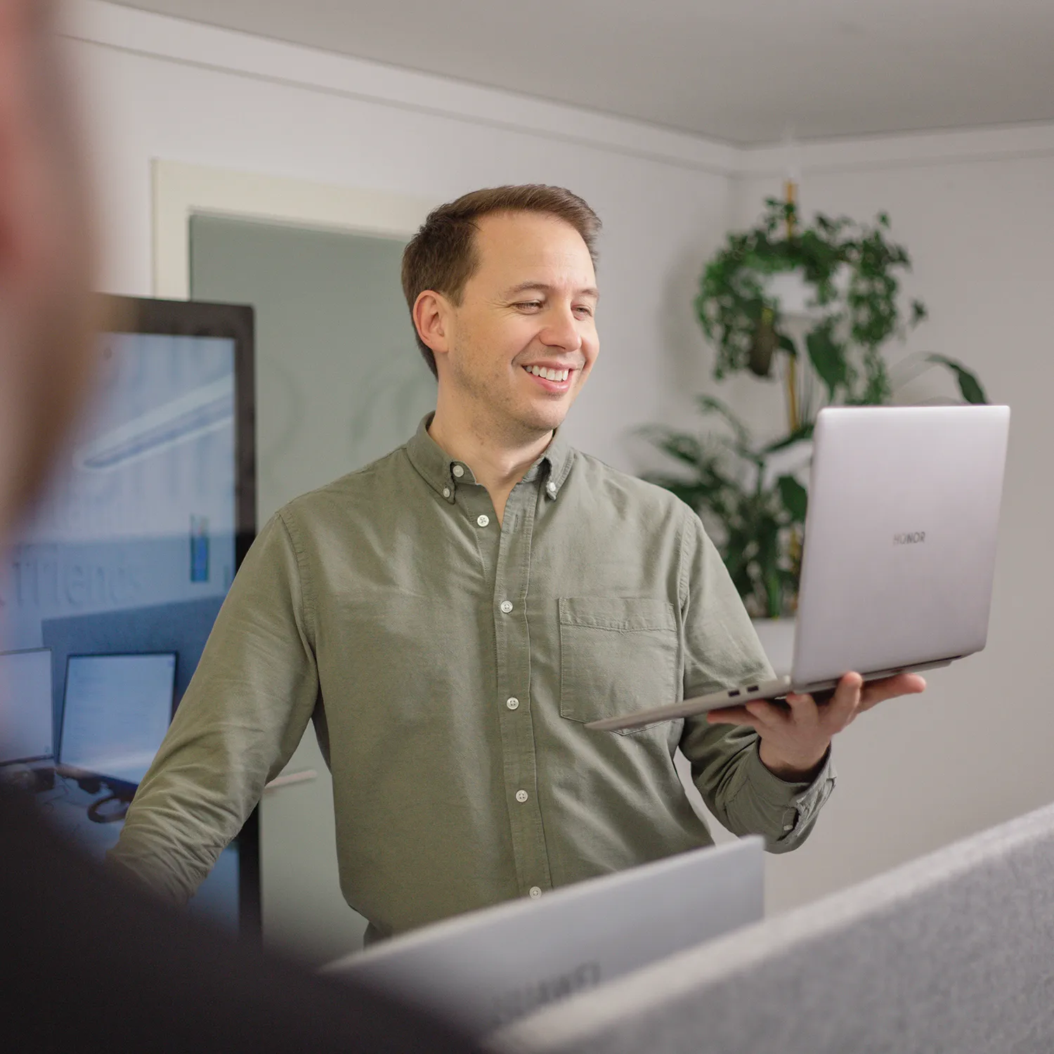 Ein Mann in einem grünen Hemd steht und lächelt, während er einen offenen Laptop in einer Büroumgebung hält, mit einer Pflanze und Computermonitoren im Hintergrund.