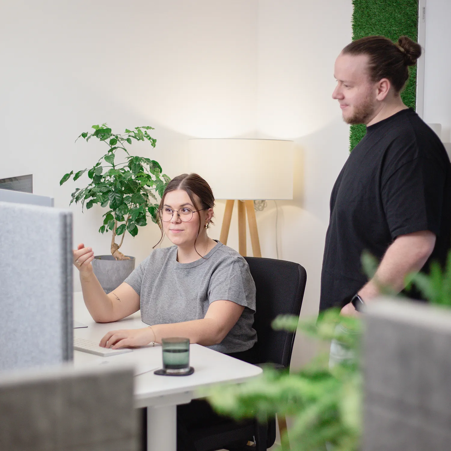 Eine Frau sitzt an einem Schreibtisch und arbeitet an einem Computer, während ein Mann neben ihr steht. Sie scheinen etwas in einem modernen Büro mit Pflanzen und einer Lampe im Hintergrund zu besprechen.