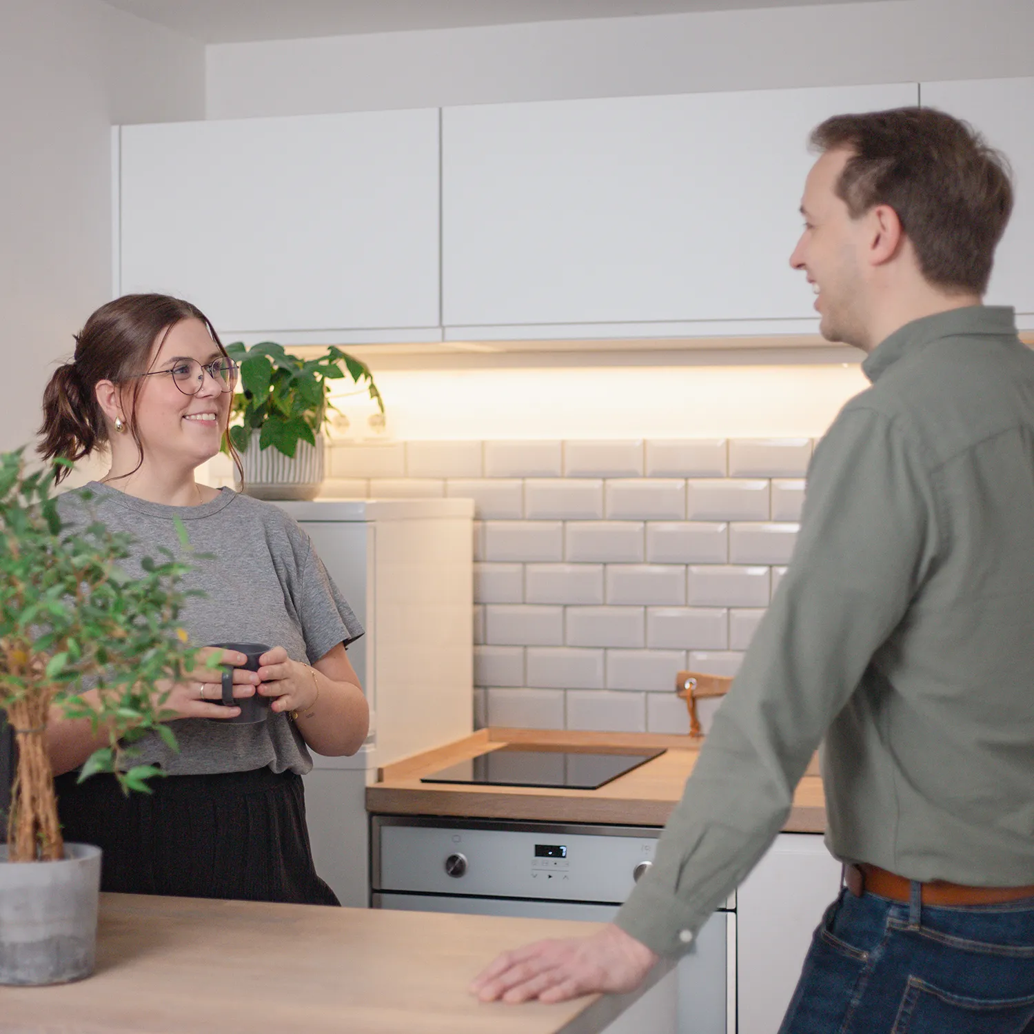 Eine Frau und ein Mann stehen in einer modernen Küche und unterhalten sich. Die Frau hält eine Tasse und lächelt. Eine Topfpflanze steht auf dem Tresen im Vordergrund.