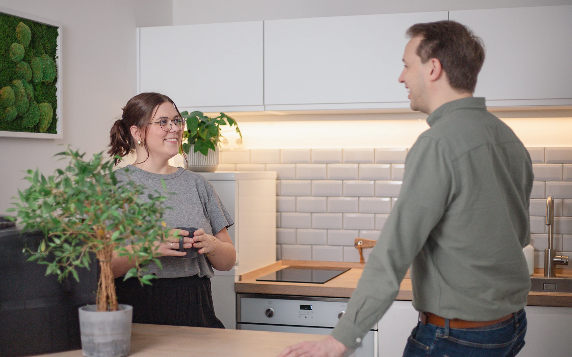 Eine Frau mit einer Tasse in der Hand und ein Mann stehen und unterhalten sich in einer modernen Küche mit weißen Schränken, gefliesten Aufkantungen und einer Topfpflanze auf dem Tresen.
