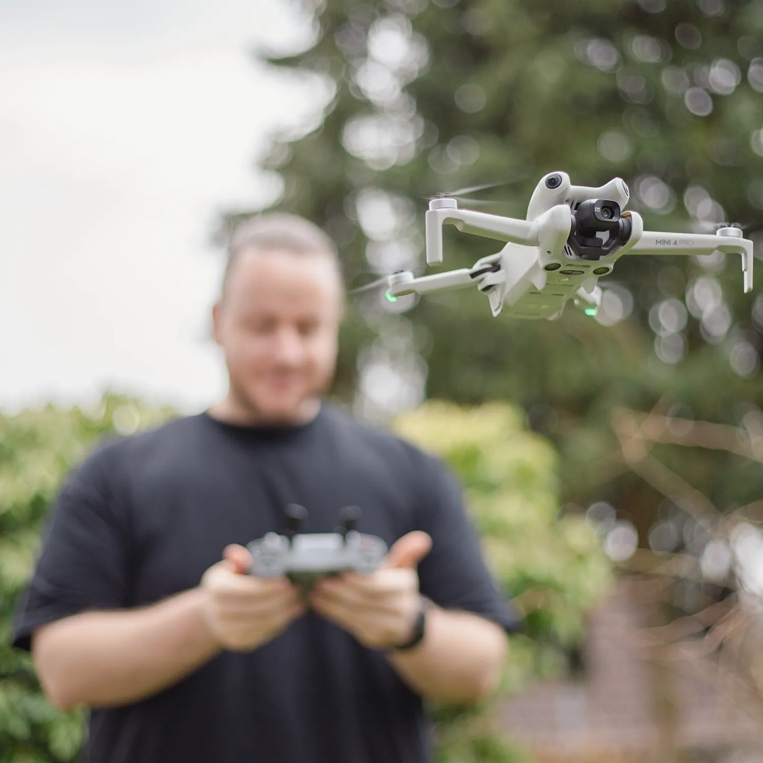 Eine Person bedient eine Fernsteuerung, während eine weiße Quadcopter-Drohne im Vordergrund im Freien schwebt.