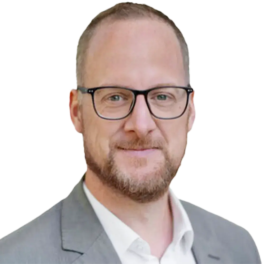 Alexander-Huhn (1) Ein Mann mit kurzen Haaren, einem Bart und einer Brille, der eine hellgraue Anzugsjacke und ein weißes Hemd trägt, lächelt leicht in die Kamera vor einem weißen Hintergrund.