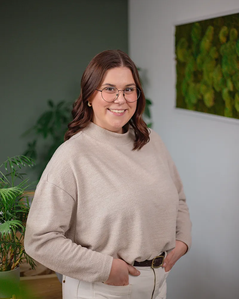 Celia Müller - Mediengestaltung Eine Frau mit schulterlangen braunen Haaren, Brille und beigefarbenem Pullover steht lächelnd in einem Haus mit Grünpflanzen und einem Bilderrahmen im Hintergrund.