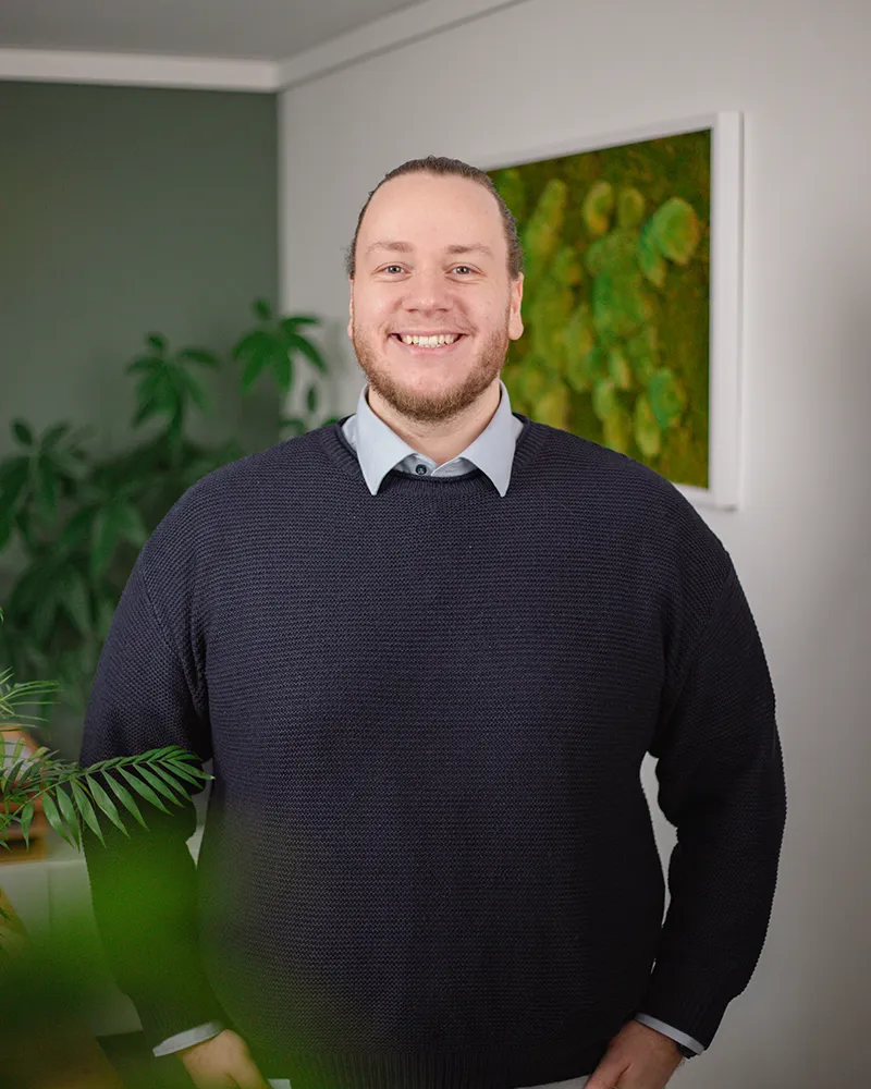 Daniel Lenz - Performance Marketing Eine Person mit heller Haut und Bart, die einen dunklen Pullover über einem Kragenhemd trägt, steht in einem Innenraum vor Grünpflanzen und einem Wandkunstwerk und lächelt in die Kamera.