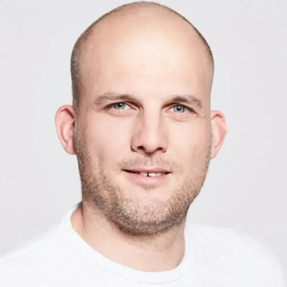 Hoffmann-Pano (1) Ein kahlköpfiger Mann mit heller Haut, blauen Augen und einem kurzen Bart blickt in die Kamera und lächelt leicht. Er trägt ein schlichtes weißes Hemd vor einem neutralen Hintergrund.