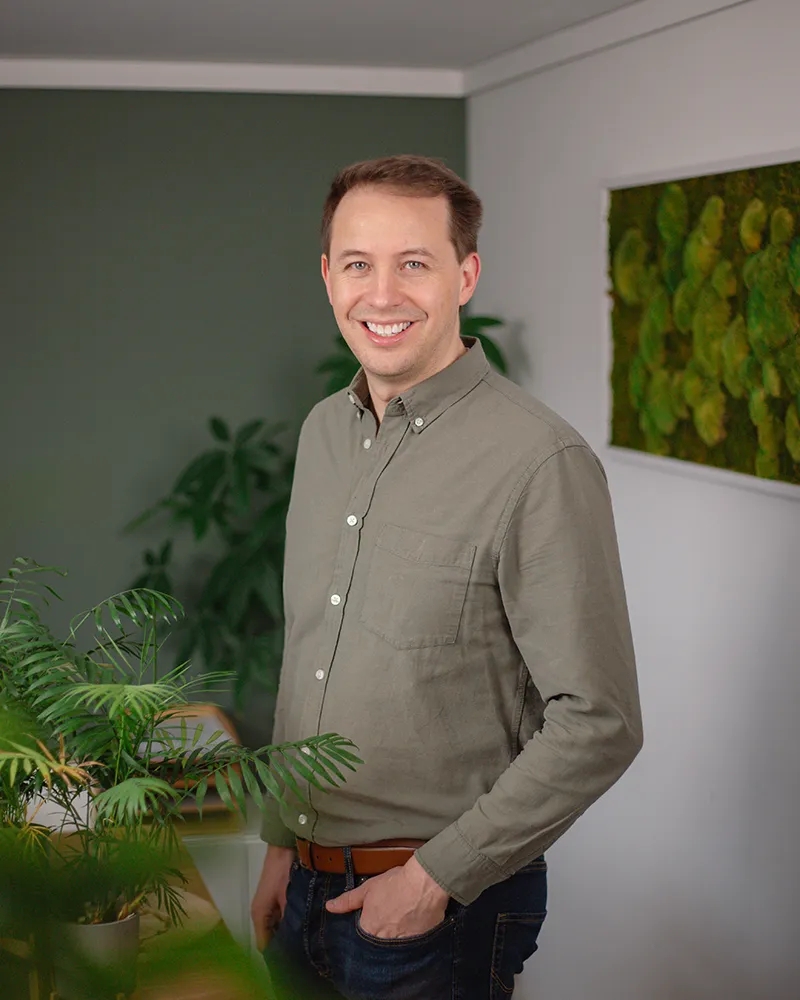 Micha Vorländer - Inhaber von Vorländer und friends Ein Mann steht in einem Haus und lächelt in die Kamera. Er trägt ein graues Button-up-Hemd und Jeans. Im Hintergrund sind Grünpflanzen und eine Wanddekoration aus Moos zu sehen.