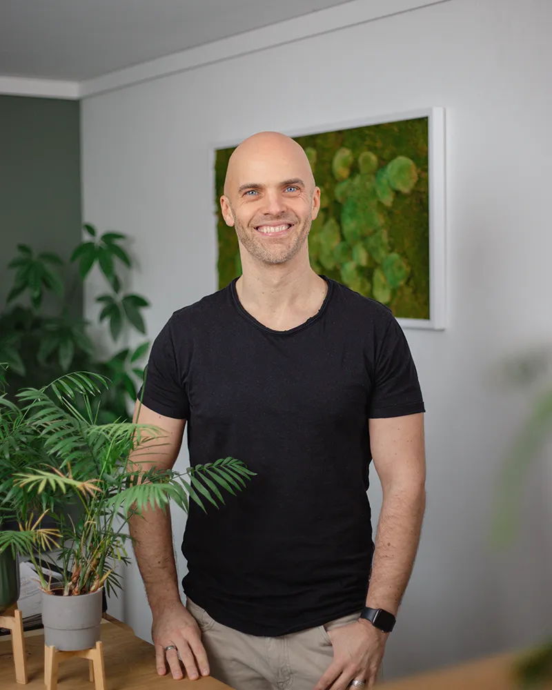 Norbert Klotz - Marketing- und Strategieberatung Ein lächelnder Mann mit Glatze und schwarzem T-Shirt steht in einem Haus in der Nähe von Topfpflanzen, mit einem grünen Wandkunstwerk im Hintergrund.