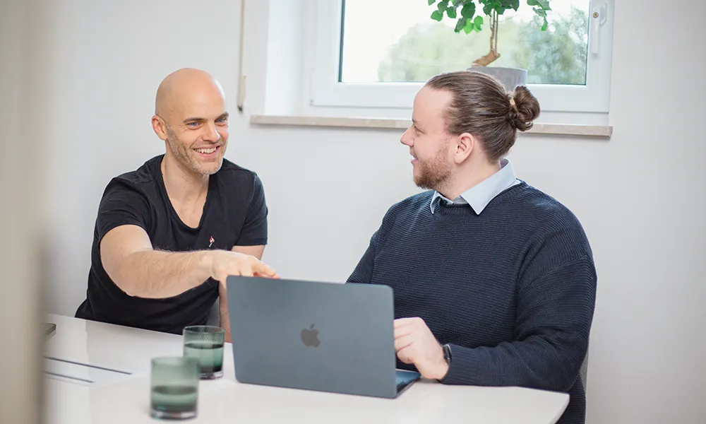 Norbert Klotz und Daniel Lenz im Meeting Zwei Männer sitzen an einem Tisch, einer mit einem Laptop, unterhalten sich und lächeln; zwei Gläser und eine Pflanze sind ebenfalls zu sehen.