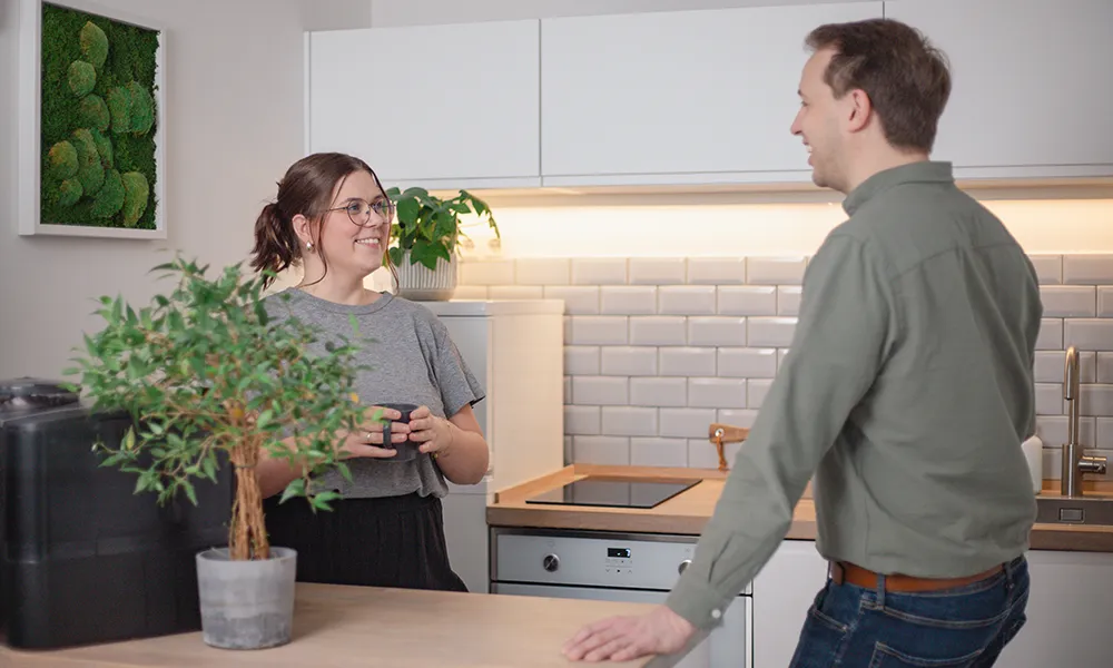 Eine Frau und ein Mann unterhalten sich in einer modernen Küche mit weißen Schränken, einer Topfpflanze und einer Kaffeemaschine auf dem Tresen.