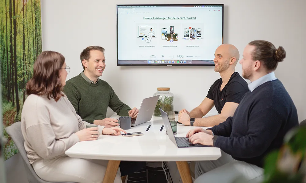 Vier Personen sitzen mit Laptops um einen Tisch herum und halten eine Besprechung in einem modernen Büro ab; auf einem Bildschirm im Hintergrund wird eine Präsentation gezeigt.