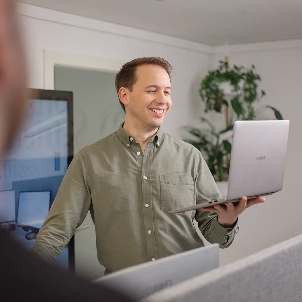 Ein Mann in einem grünen Hemd steht und lächelt, während er einen offenen Laptop in einer Büroumgebung hält, mit Pflanzen und Computermonitoren im Hintergrund.