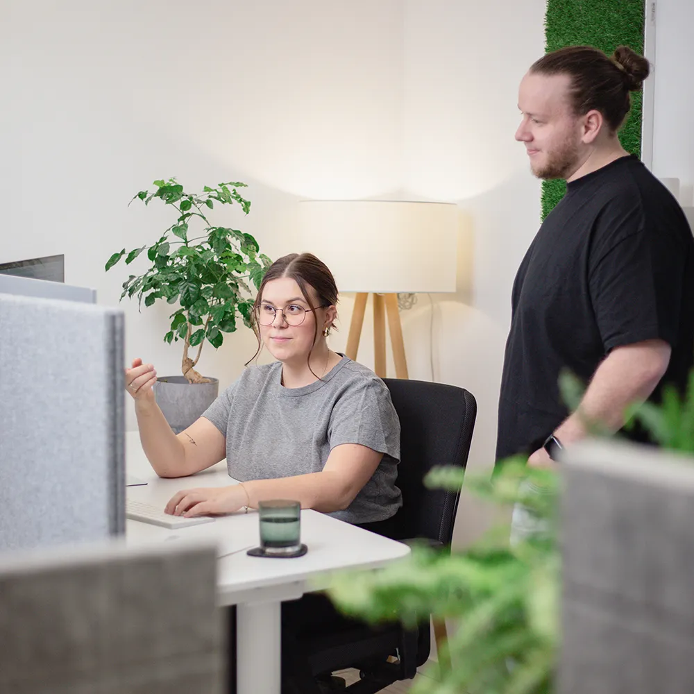 Eine Frau sitzt an einem Schreibtisch mit einem Computer und spricht gestikulierend mit einem Mann, der neben ihr in einem modernen Büroraum mit Pflanzen und einer Lampe steht.