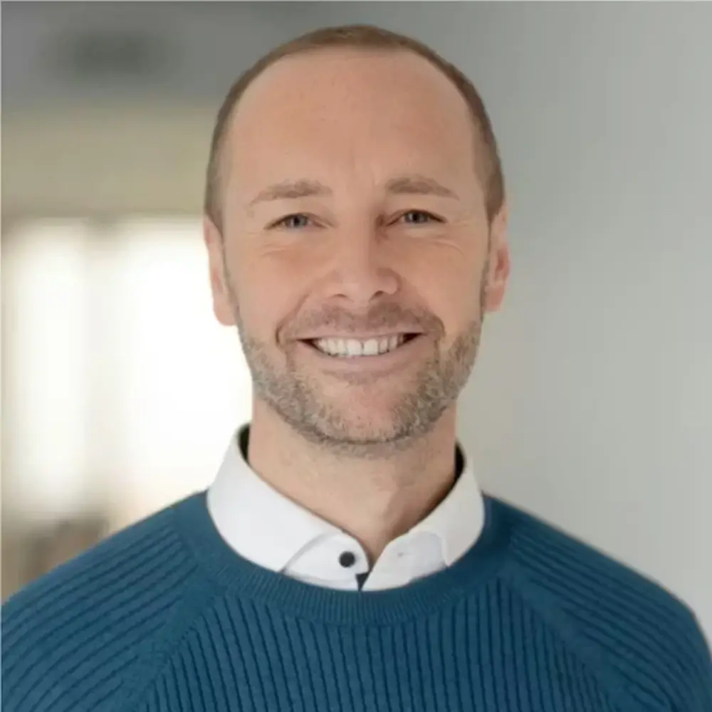 alexander herholz (1) Ein Mann mit kurzen Haaren und gestutztem Bart lächelt, trägt einen blauen Pullover über einem weißen Hemd mit Kragen und steht in einem Haus mit unscharfem Hintergrund.