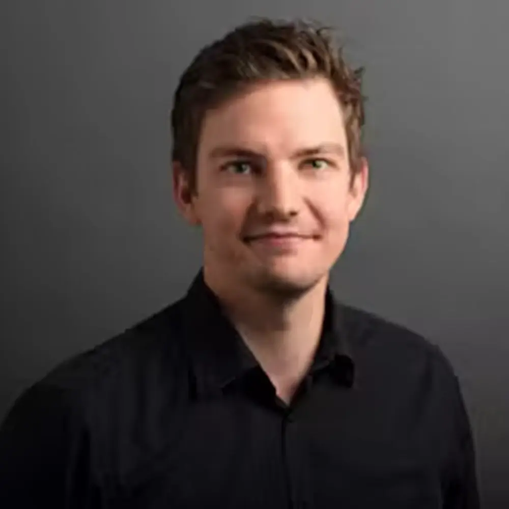 eike eschmann (1) Ein Mann mit kurzen braunen Haaren und schwarzem Kragenhemd steht vor einem schlichten grauen Hintergrund und blickt mit einem leichten Lächeln in die Kamera.