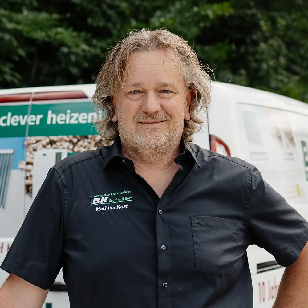 mathias kost (1) Ein Mann mit gewelltem Haar und Bart steht vor einem Lieferwagen und trägt ein dunkles kurzärmeliges Hemd mit aufgesticktem Namen und Logo. Im Hintergrund sind Bäume zu sehen.